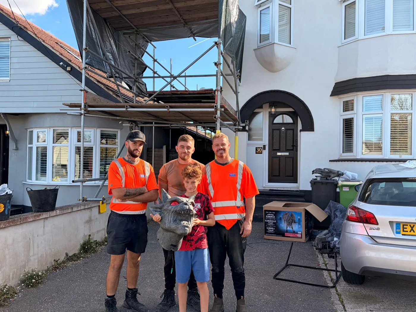 Dexter holding a gargoyle head, standing between three men from TG Scaffolding in front of the scaffolded house on Queens Road
