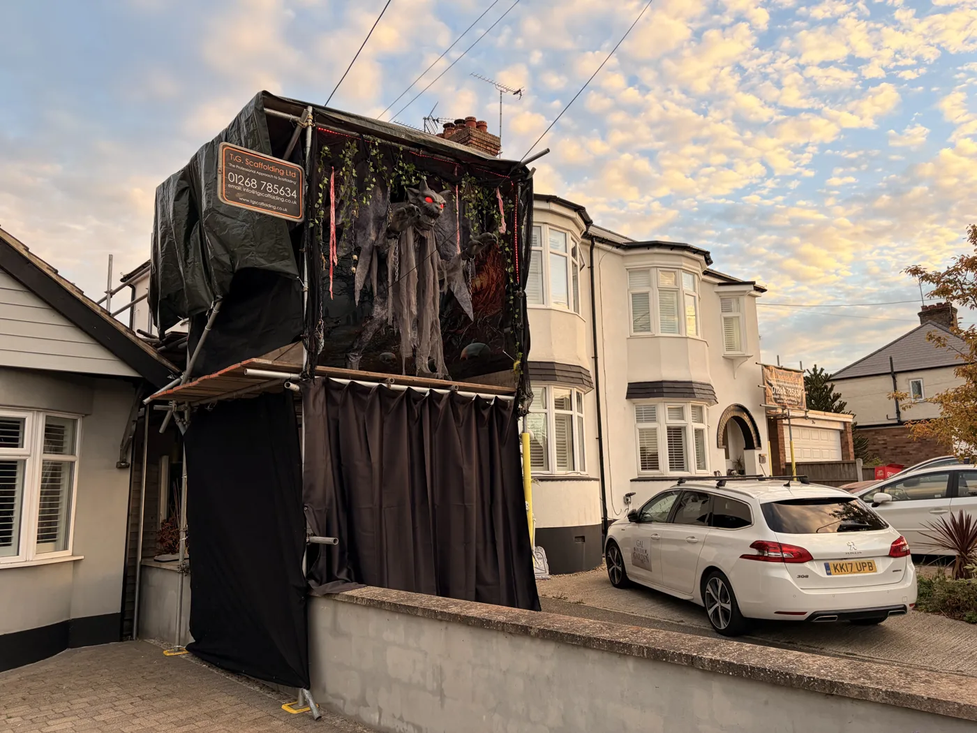 Scaffolding wrapped in black sheeting in front of a semi-detached house, with a winged gargoyle figure on a platform above a curtained entrance